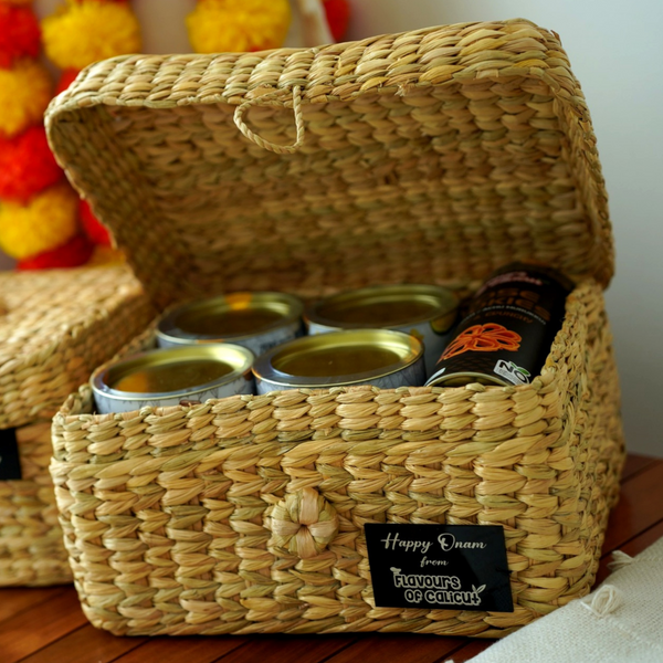 Woven basket with tins and a bottle, labeled 'Happy Onam from Flavours of Calicut', on a wooden surface.